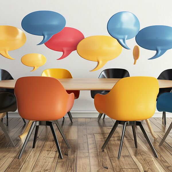 St&uuml;hle in verschiedenen Farben stehen um einen runden Tisch. &Uuml;ber dem Tisch befinden sich bunte Sprechblasen.