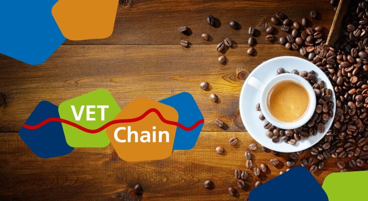 Ein Holztisch auf dem Kaffeebohnen und eine Tassee Kaffee stehen. Das Logo und einzelne bunte Elemente des Logos der VET Chain &uuml;berlagern das Foto