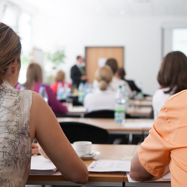 Das Bild zeigt Erwachsene in einem Schulungsraum. Die Menschen sitzen an ihren Schreibtischen und werden von hinten gezeigt wie sie auf die Tafel am Ende des raumes schauen.
